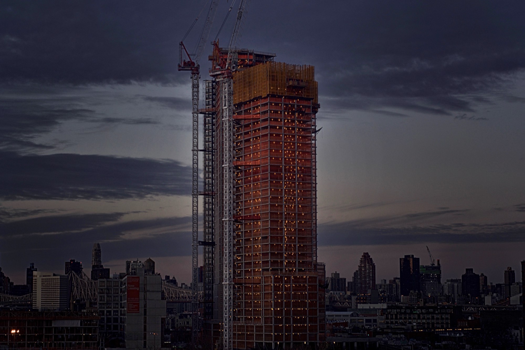 A tower under construction lit red against the New York skyline at dusk.