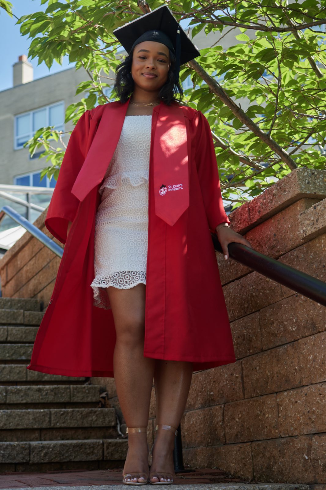 Full length on the steps in cap and gown, sunlight overhead.