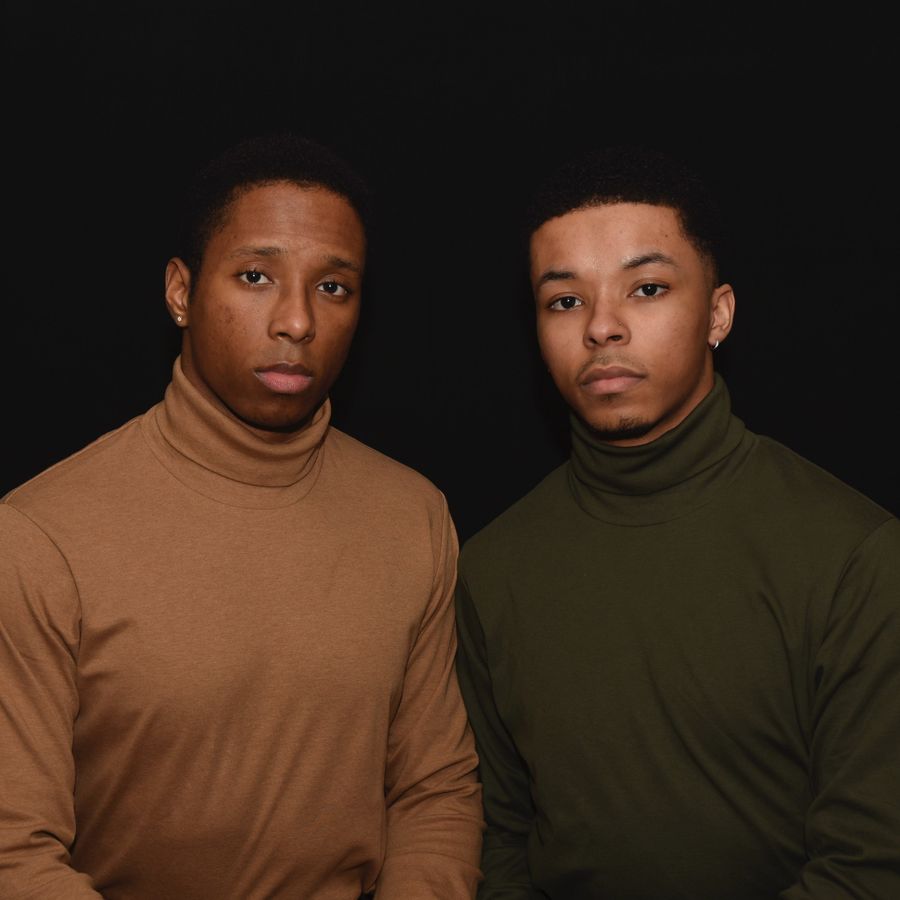Studio portrait of two men in turtlenecks, one tan and one olive, against a black backdrop.