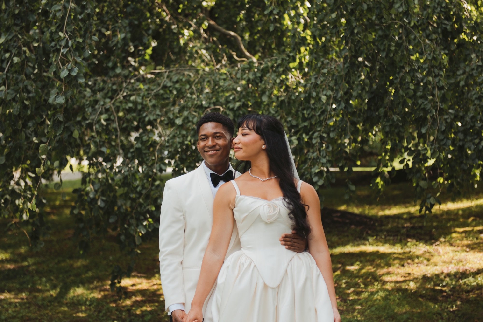 The couple standing close under a canopy of leaves, the groom smiling.