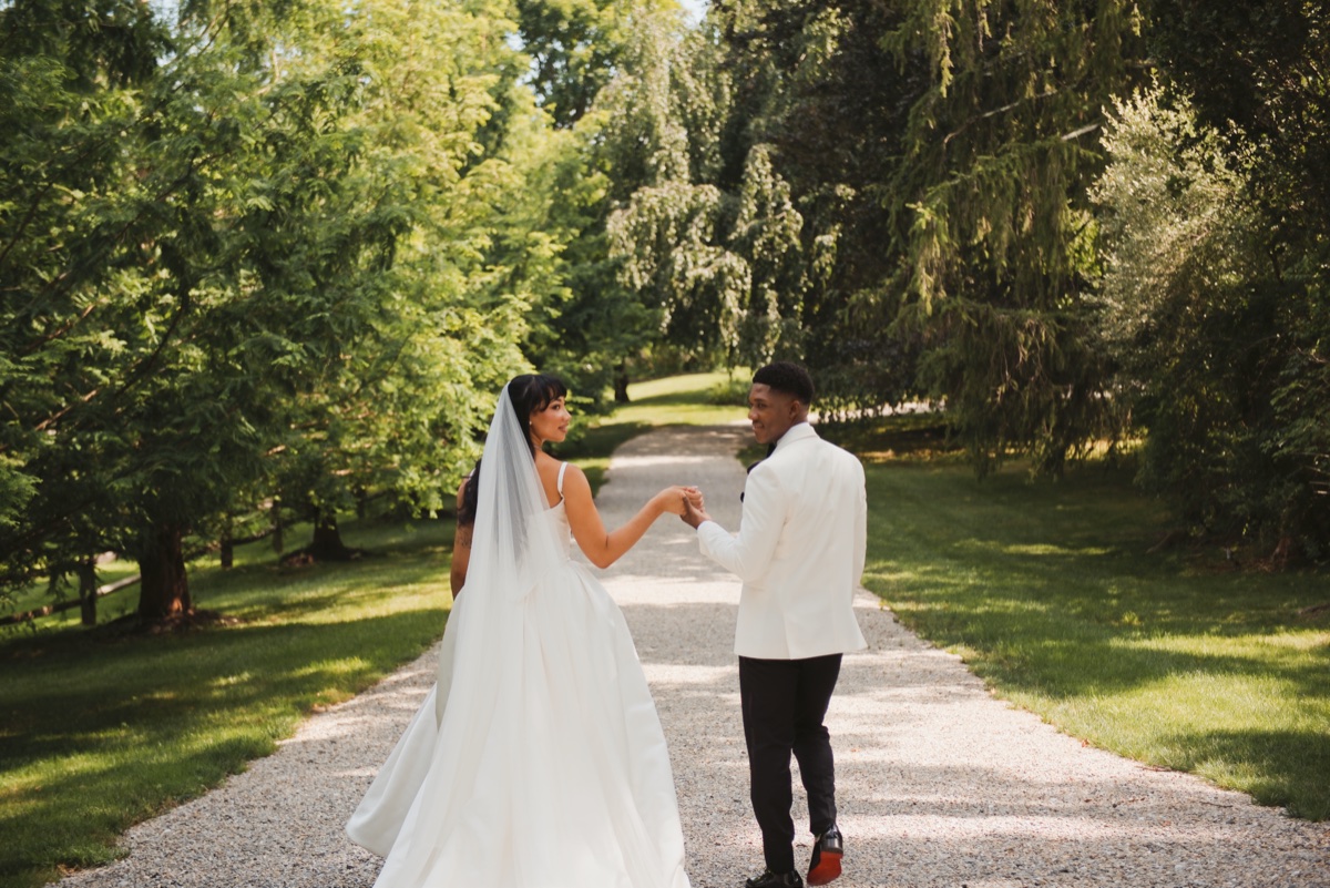 A bride and groom walking down a gravel drive between tall trees, holding hands.